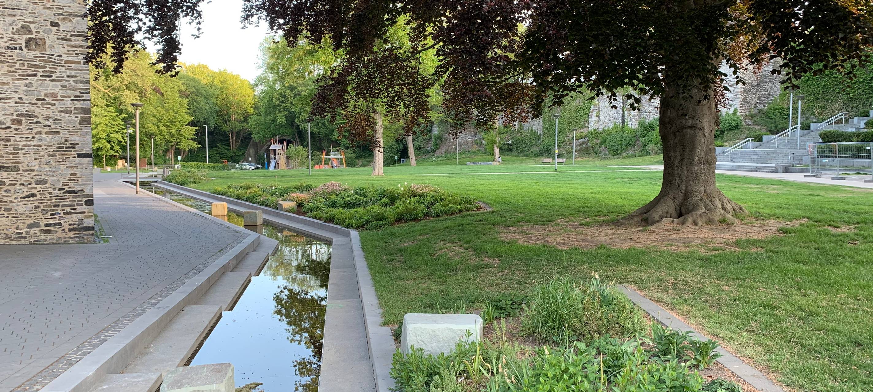 Künstlicher Bachlauf an einer Kirche auf einem grünen Platz in Iserlohn. Auf dem Platz steht eine Buche.