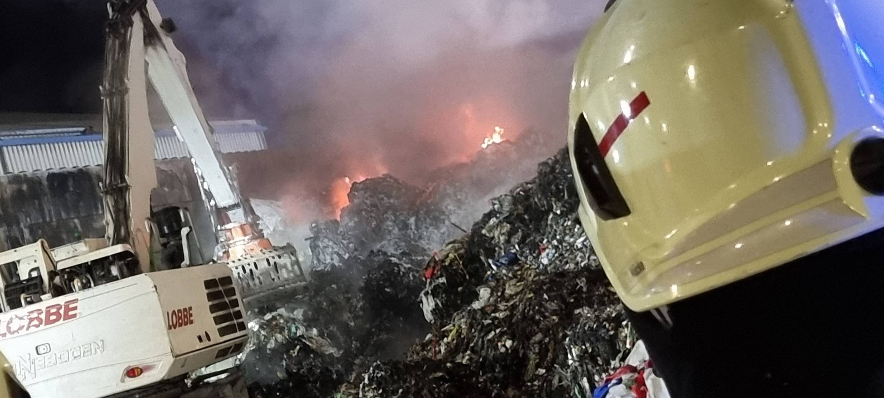 Ein Feuerwehrmann blickt auf einen großen Haufen aus verbranntem Plastikmüll. Es qualmt.