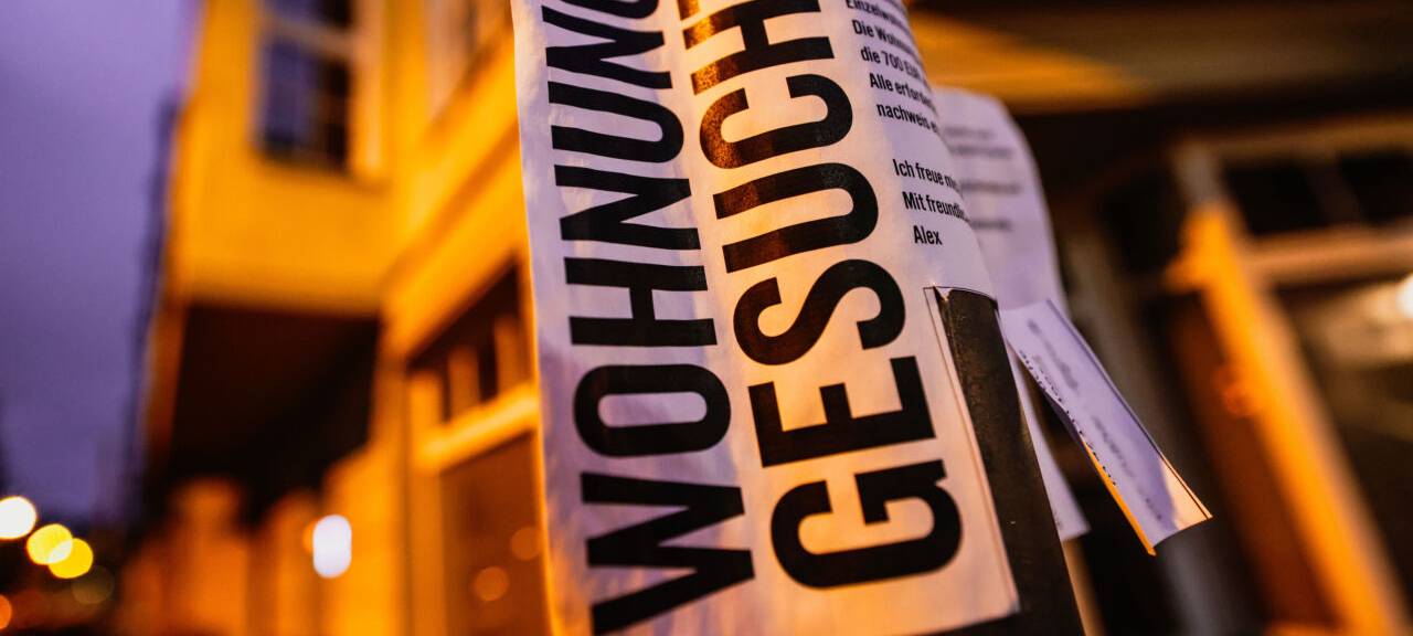 Ein Zettel mit der Aufschrift "Wohnung gesucht" hängt vor Wohnhäusern.