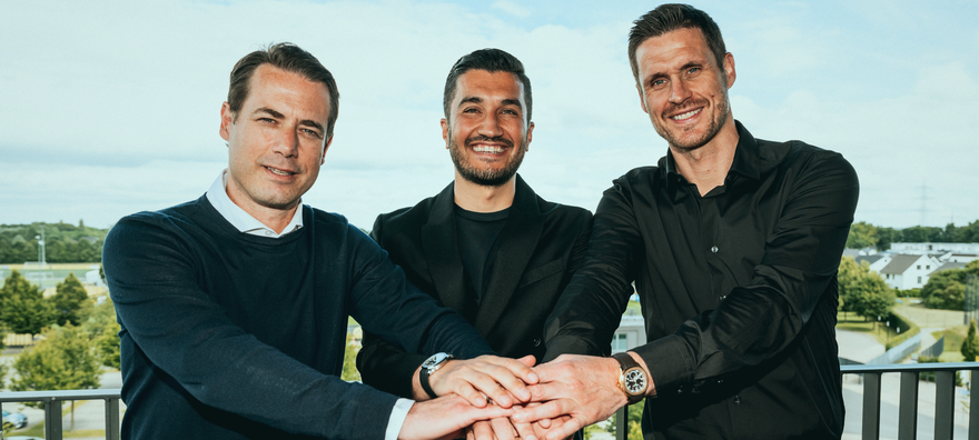 Drei Männer mit braunen Haaren halten ihre Hände zusammen. Sie lachen in die Kamera.